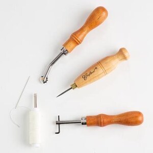 May include: A set of three leather sewing tools with wooden handles. The tools include a stitching awl, a stitching groover, and a stitching punch. A spool of white waxed thread and a needle are also included.