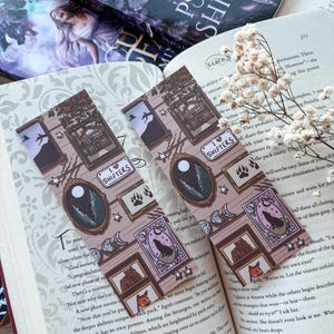 May include: Two decorative bookmarks with a brown and cream colour scheme, featuring framed illustrations of a house, a wolf, a bear, and a fox. The bookmarks are placed on an open book with text and a sprig of white flowers.