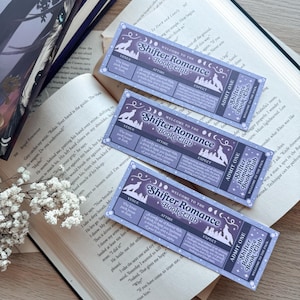 May include: Three purple and white Shifter Romance Book Club tickets are displayed on an open book. The tickets feature a wolf silhouette, moon phases, and event details. Books are in the background.