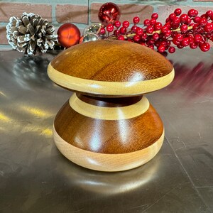 May include: A wooden mushroom-shaped decorative object with a brown and light wood finish. The top of the mushroom is a flat, round surface, and the bottom is a rounded, bulbous shape. The object is made of multiple pieces of wood that are joined together to create a striped pattern.