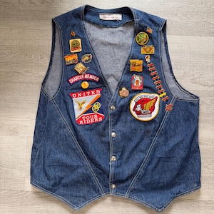 May include: A vintage denim vest with multiple patches and pins. Patches include "Charter Member," "United Tour Riders," and "Wings." The vest is button-down with a V-neck and a faded blue color. A classic piece of apparel.