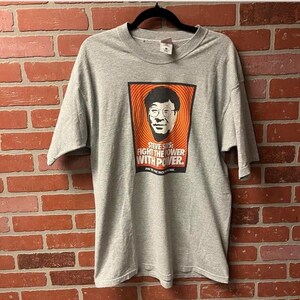 May include: A gray t-shirt with a graphic featuring a portrait of a man with glasses against an orange and red background. The text "STEVE SAYS: FIGHT THE POWER WITH POWER" is printed below the portrait.
