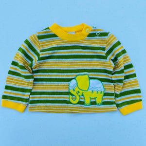 May include: A long-sleeved baby shirt with horizontal stripes in green, yellow, and white. The shirt features a yellow collar and cuffs, and a yellow elephant applique with white accents on the front. The shirt has a button closure at the shoulder.