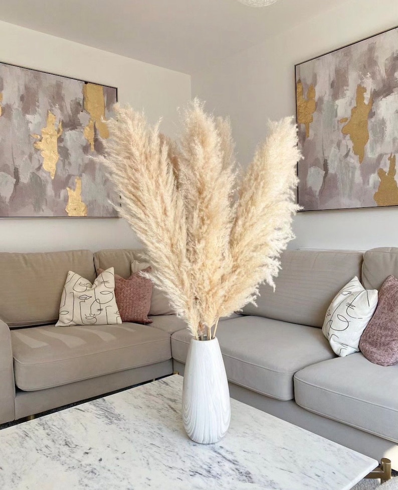 May include: A white vase with a textured surface holds a bouquet of dried pampas grass. The vase is on a white marble coffee table in front of a gray couch with pillows. Two abstract paintings with gold accents hang on the wall behind the couch.