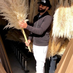 May include: A person holding a large bundle of dried pampas grass. The grass is a light beige color and is arranged in a fan shape. The person is wearing a black sweatshirt and white pants. The background is a warehouse with boxes and other dried pampas grass.