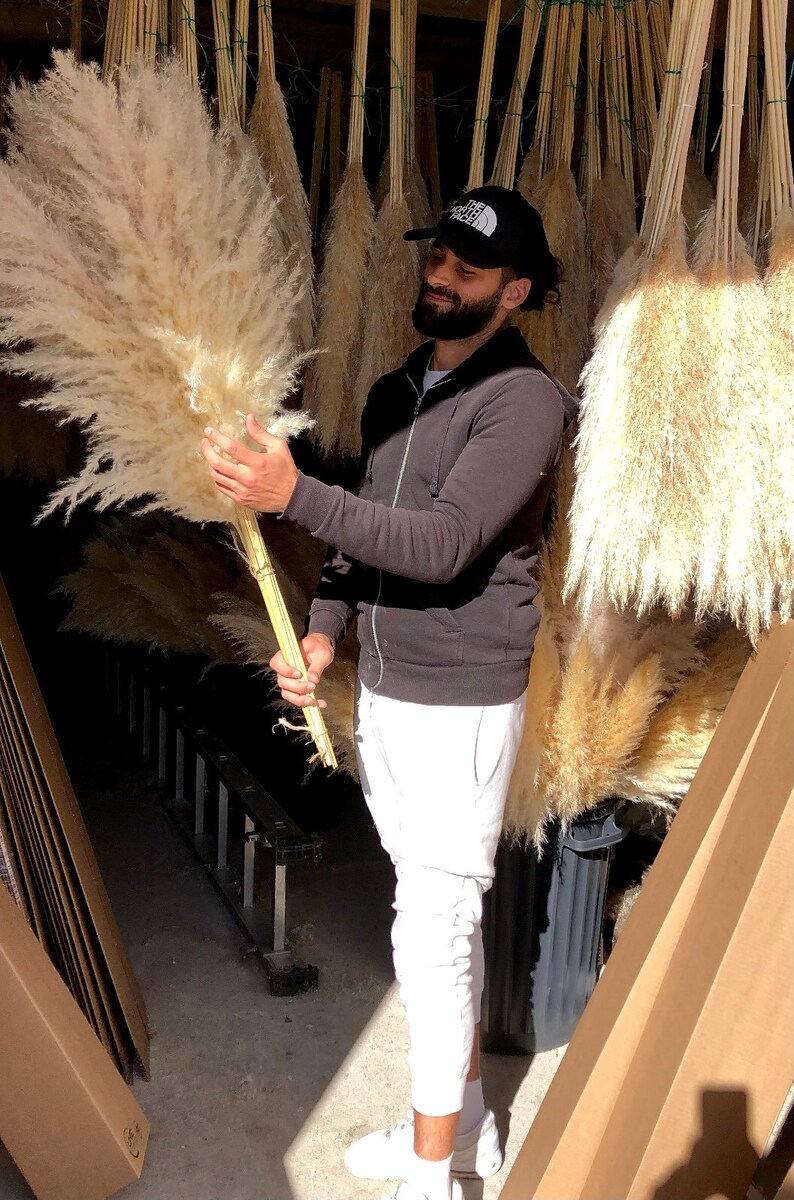 May include: A person holding a large bundle of dried pampas grass. The grass is a light beige color and is arranged in a fan shape. The person is wearing a black sweatshirt and white pants. The background is a warehouse with boxes and other dried pampas grass.