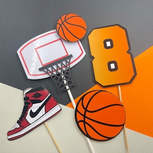 May include: A set of five basketball-themed cake toppers. The toppers include a basketball hoop, a basketball, a basketball shoe, the number eight, and another basketball. The toppers are all orange, black, and white.