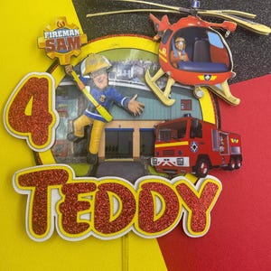 May include: Fireman Sam themed cake topper featuring a red helicopter, fire engine, and a figure of Fireman Sam. The topper also includes a red glitter number 4 and the name "Teddy" in red glitter.