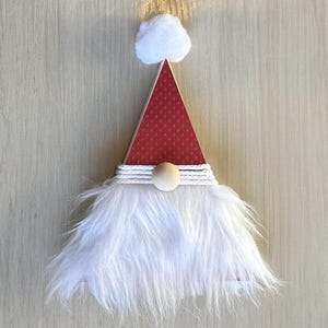 May include: A wooden gnome Christmas ornament. The red hat has a dotted pattern and a white pom-pom. The beard is made of fluffy white material, and the hat is wrapped with white twine. A wooden ball is in the center.