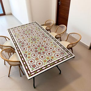 Peut inclure: Une table à manger rectangulaire dotée d'un plateau blanc orné d'un motif géométrique complexe en mosaïque dans des tons de rouge, vert et gris, reposant sur des pieds en métal noir, entourée de six chaises en osier clair à coussins blancs.
