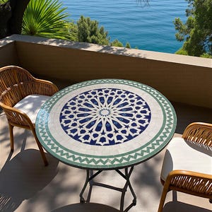 Peut inclure: Une table ronde en mosaïque avec un motif d'étoile géométrique bleu et blanc entouré d'une bordure en zigzag verte, sur un cadre en métal, accompagnée de deux chaises en osier marron clair avec des coussins blancs.
