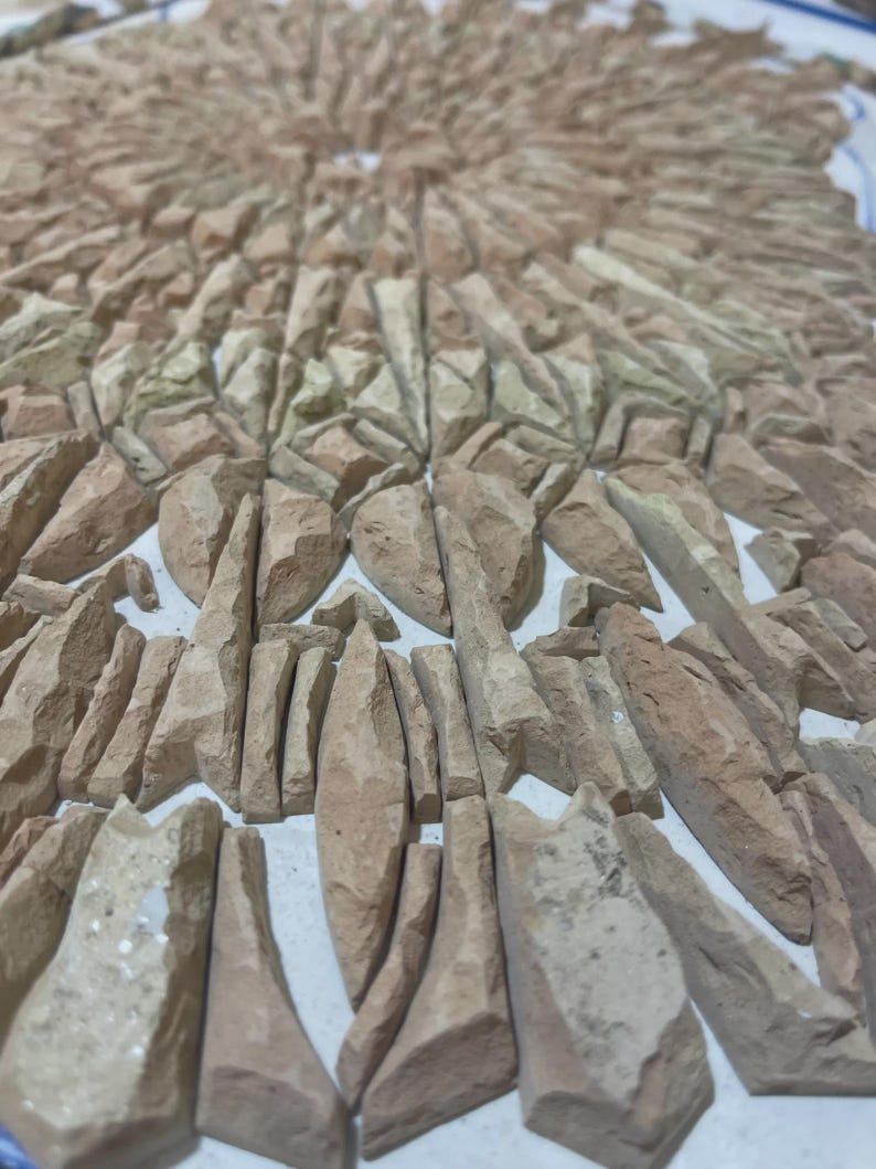 May include: A mosaic of irregularly shaped, light brown stones arranged in a radial pattern. The stones vary in size and shape, creating a textured surface. The arrangement resembles a sunburst design, with the stones radiating outwards from a central point.