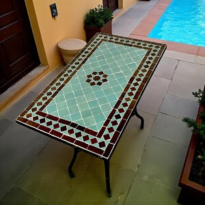 May include: A rectangular outdoor table with a mosaic tile top. The table has a light teal centre with a brown starburst design and a brown and red diamond border. The table has black metal legs.