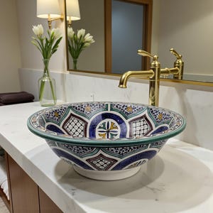 May include: A patterned ceramic sink with a detailed design in blue, green, and white. The sink is set against a backdrop of a gold tap, mirror, and lighting. The sink's design includes geometric shapes and patterns.