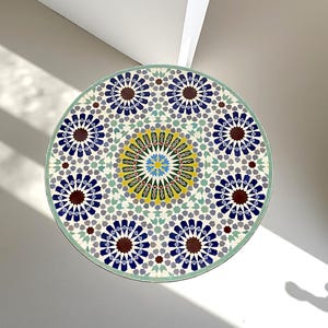 May include: A circular table top featuring a complex geometric mosaic pattern. The design includes a central star motif in yellow, green, and blue, surrounded by repeating dark blue and brown star-shaped tiles on a white and light green background.