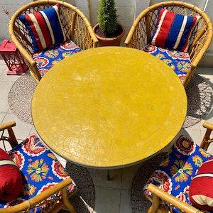 Pode incluir: Uma mesa redonda com tampo de mosaico amarelo rodeada por quatro cadeiras de vime. Cada cadeira possui uma almofada de assento azul com um padrão floral colorido e uma almofada retangular com listras verticais vermelhas, brancas e azuis.