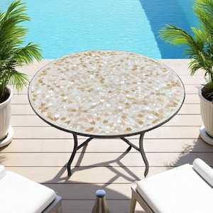 May include: A round outdoor table featuring a mosaic tile top with a geometric pattern in shades of white, cream, and tan. The table is supported by a black metal frame with four curved legs that meet in a central point beneath the tabletop.
