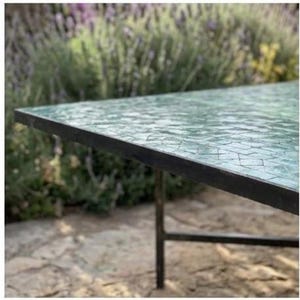 May include: A rectangular table with a mosaic glass top in shades of green and blue. The table has a black metal frame and legs. The table is outdoors, with a blurred background of greenery and stone.
