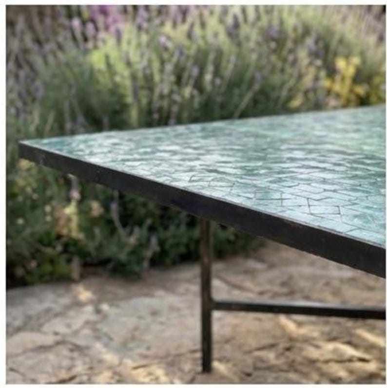 May include: A rectangular table with a mosaic glass top in shades of green and blue. The table has a black metal frame and legs. The table is outdoors, with a blurred background of greenery and stone.