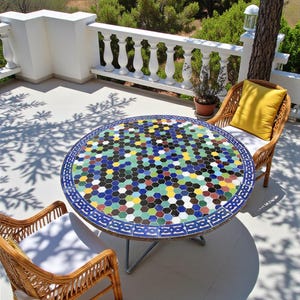 Könnte beinhalten: Runder Gartentisch mit Mosaik-Tischplatte mit farbenfrohem geometrischem Muster. Zwei Korbstühle mit weißen Kissen und einem gelben Kissen umgeben den Tisch. Die Szene befindet sich auf einer Terrasse mit weißem Geländer und viel Grün im Hintergrund. Durchmesser ca. 100 cm.