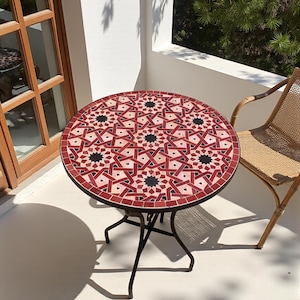 Może przedstawiać: Okrągły stół zewnętrzny z blatem z mozaiki. Blat ma geometryczny wzór w kolorach czerwonym, czarnym i kremowym. Stół ma ciemną metalową ramę i ma średnicę około 61 cm.