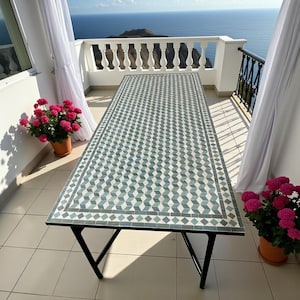 May include: A rectangular outdoor table with a mosaic tile top featuring a geometric cube pattern in shades of teal, gray, and white. The table has black metal legs. Potted pink flowers and a scenic ocean view are in the background.