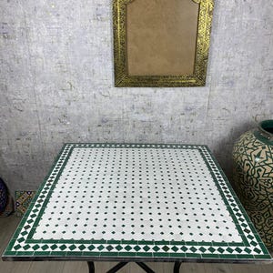 Peut inclure: Une table carrée dotée d'un plateau carrelé blanc avec un motif répétitif de petits losanges verts. Le plateau est bordé d'un cadre rectangulaire vert épais et repose sur une base en métal noir munie de quatre pieds fins.