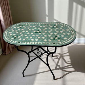 Peut inclure: Une table ovale dotée d'un plateau en mosaïque avec un motif répétitif d'étoiles blanches et vert clair, accentué par des motifs triangulaires vert foncé. Le plateau est bordé d'un motif en losanges et repose sur une base en métal noir.