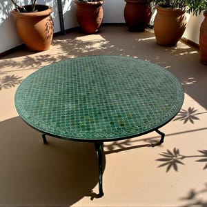 May include: A round coffee table featuring a green mosaic tile top with a geometric pattern and a thin black metal border. The table is supported by three slender black metal legs that curve outward, providing a stable base for the circular surface.