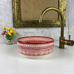 Puede incluir: Un cuenco de cerámica decorativo con borde rojo y un patrón geométrico blanco y rojo. El cuenco está sobre una superficie blanca, junto a un grifo de latón y un pequeño jarrón de flores.