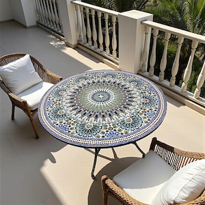 May include: A round table with a blue, green, and white geometric mosaic tile top and a metal base, accompanied by two woven wicker chairs featuring white seat cushions and back pillows, arranged on a balcony with a white balustrade railing.