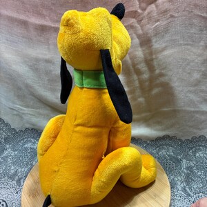 Op de afbeelding: Een gele Pluto-knuffel met zwarte oren en staart en een groene halsband. Het speelgoed zit op een houten oppervlak. Een populair Disney-personage, gemaakt van zacht materiaal.