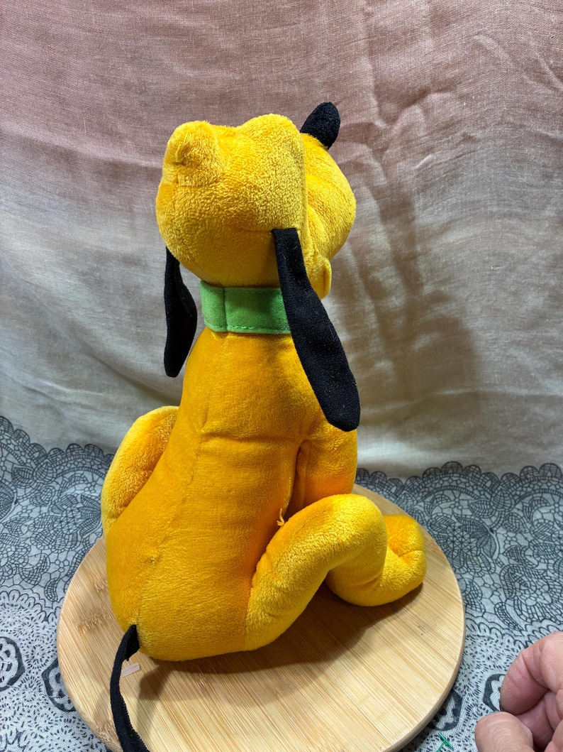 Op de afbeelding: Een gele Pluto-knuffel met zwarte oren en staart en een groene halsband. Het speelgoed zit op een houten oppervlak. Een populair Disney-personage, gemaakt van zacht materiaal.