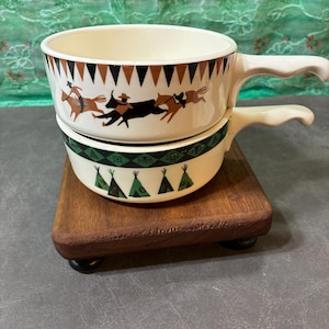 May include: Two cream-colored soup bowls with handles, stacked on a wooden stand. The top bowl features a brown and black geometric pattern and a scene of riders and animals. The bottom bowl has a green and black geometric pattern.