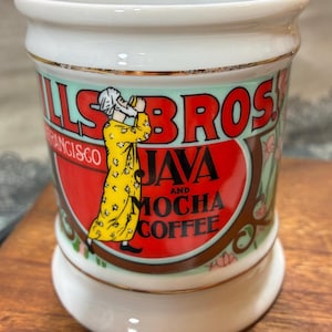 May include: A white ceramic coffee mug with gold trim. The mug features a red and green label with the words "ILLS BROS JAVA AND MOCHA COFFEE" and an illustration of a man in a yellow coat.