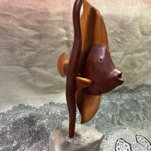 May include: A wooden sculpture of a fish with a dark brown body and a lighter brown fin. The fish is mounted on a small, square, gray stone base. The sculpture has a smooth finish and is likely handmade.