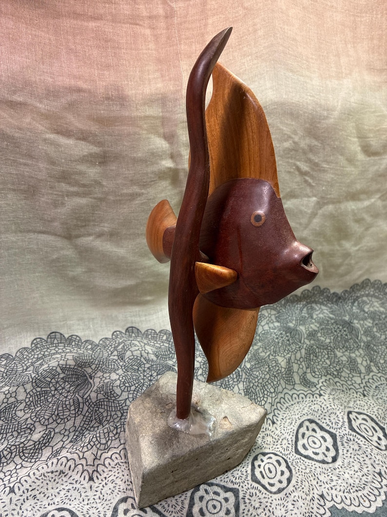 May include: A wooden sculpture of a fish with a dark brown body and a lighter brown fin. The fish is mounted on a small, square, gray stone base. The sculpture has a smooth finish and is likely handmade.