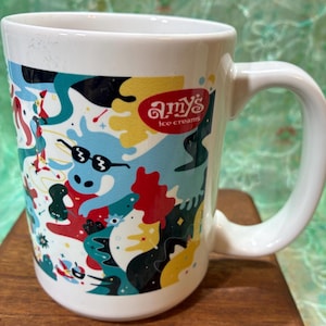May include: White ceramic mug with a wraparound design featuring a whimsical illustration with a blue hippo wearing sunglasses, and the text "Amy's ice creams". The design includes red, yellow, and black abstract shapes.