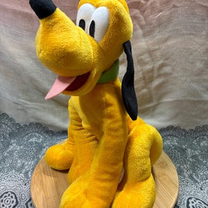 Op de afbeelding: Een pluche speelgoed van Pluto, een gele hond met zwarte oren en neus, een groene kraag en een roze tong. Het speelgoed staat op een ronde houten basis.