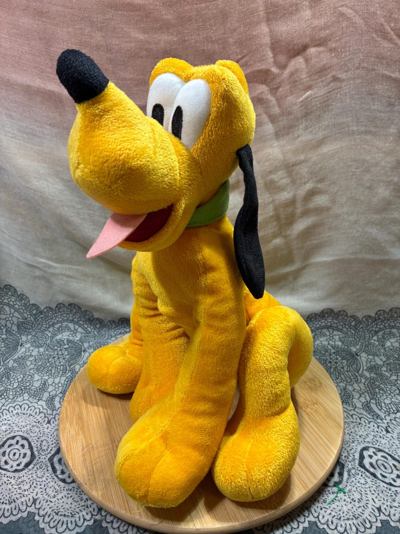 Op de afbeelding: Een pluche speelgoed van Pluto, een gele hond met zwarte oren en neus, een groene kraag en een roze tong. Het speelgoed staat op een ronde houten basis.