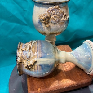 May include: Two handmade ceramic wine goblets. The goblets are glazed in shades of blue, cream, and brown. Each goblet features a raised relief design of grape leaves and grapes. The stem and base are a lighter cream color.