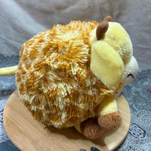 May include: A plush toy Highland cow with a fluffy, orange and yellow body. The cow has yellow ears, brown horns, and brown hooves. It is sitting on a round wooden plate.