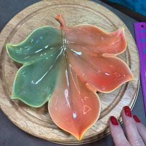 May include: A ceramic leaf-shaped dish with a glossy finish. The dish is divided into two sections, one green and the other orange. The dish is placed on a wooden surface.