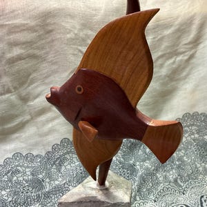 May include: A wooden fish sculpture with a dark brown body and light brown fins. The sculpture is mounted on a gray stone base. The fish has a detailed design and is a decorative item.