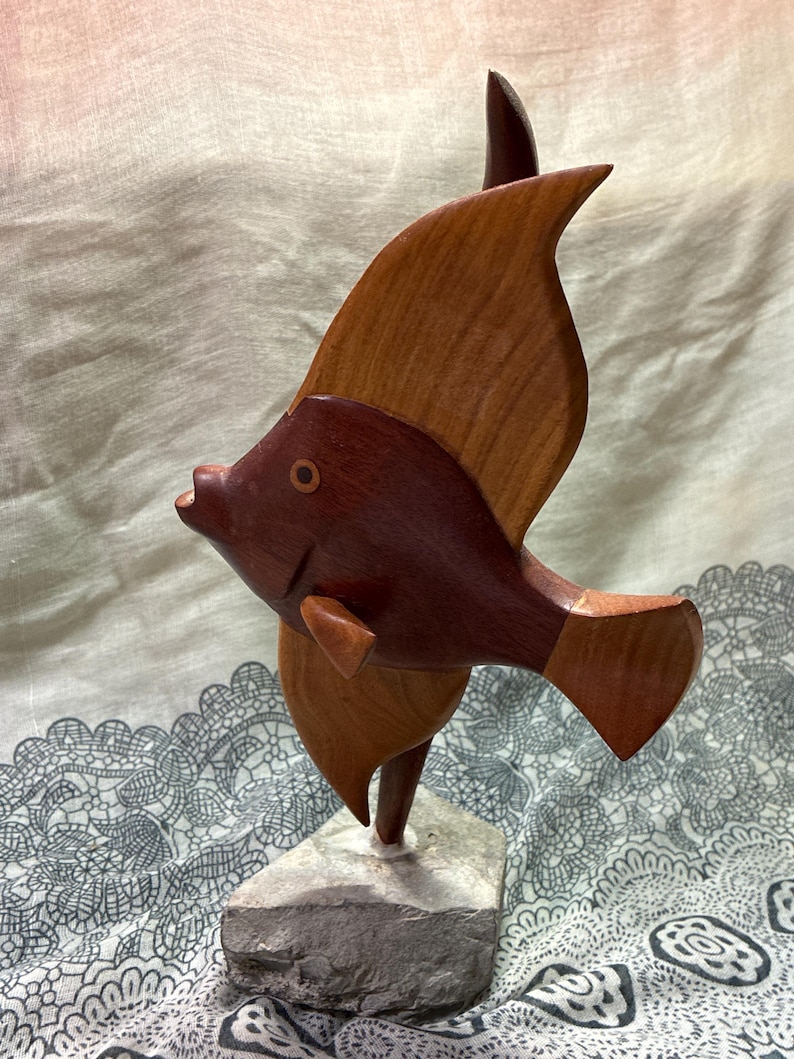 May include: A wooden fish sculpture with a dark brown body and light brown fins. The sculpture is mounted on a gray stone base. The fish has a detailed design and is a decorative item.