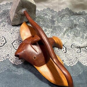 May include: A handcrafted wooden sculpture of a fish with a dark brown head and a lighter brown body. A dark brown curved piece extends from the fish, connecting to a gray stone. The sculpture is set against a patterned background.