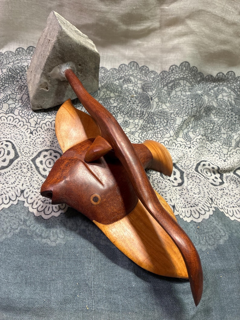 May include: A handcrafted wooden sculpture of a fish with a dark brown head and a lighter brown body. A dark brown curved piece extends from the fish, connecting to a gray stone. The sculpture is set against a patterned background.