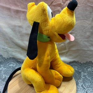 Op de afbeelding: Een gele Pluto-knuffel met zwarte oren en neus. Het heeft een groene kraag en een roze tong. Het speelgoed zit op een houten oppervlak.