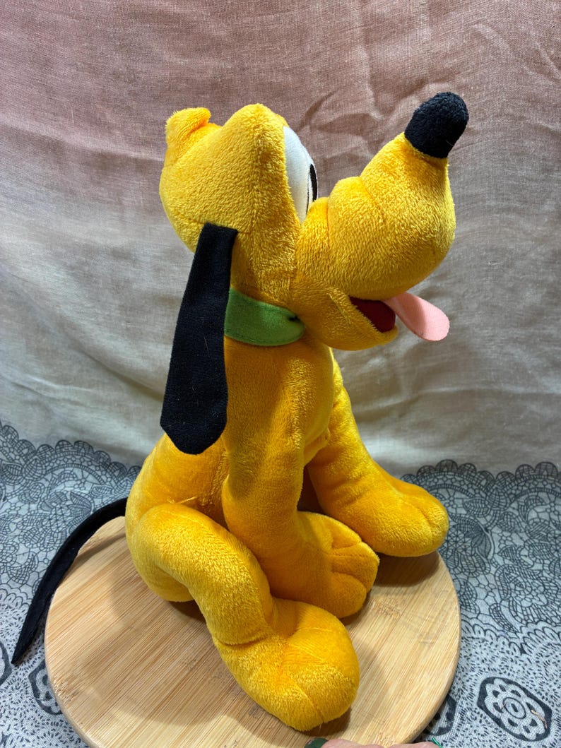 Op de afbeelding: Een gele Pluto-knuffel met zwarte oren en neus. Het heeft een groene kraag en een roze tong. Het speelgoed zit op een houten oppervlak.
