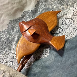 May include: A wooden fish sculpture with a detailed design. The fish is crafted from two shades of wood, with a darker brown head and a lighter brown body and fins. The sculpture has a whimsical, handcrafted appearance.